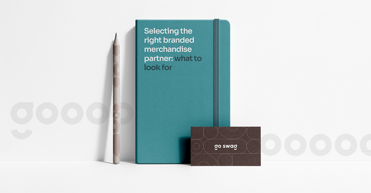 Branded merchandise flat lay featuring a teal notebook, matching pen, and Go Swag business card, illustrating how to choose the right branded merchandise partner.